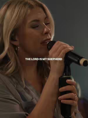 A medium shot of a woman with blonde hair, illuminated from the front, holding a microphone in her right hand while singing. The background is blurred, suggesting a performance setting.