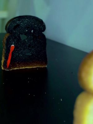 Image shows a close-up of a charred piece of toast with a streak of red paint that looks like blood. It is on a dark surface, possibly a table, with a blurry, golden object to the right, a light blue wall is in the background, with various small, white specks scattered on the dark surface.