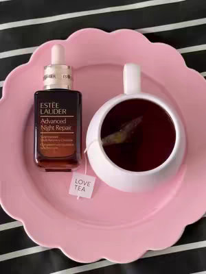 A close-up, top-down shot featuring a dark brown bottle of Estée Lauder Advanced Night Repair serum, a white teacup with dark tea and a tea bag, and a pink scalloped plate, all set against a black and white striped background.