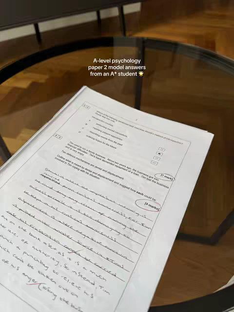 A photo of a student's A-level psychology exam paper and annotations. The paper is open on a wooden table, with the text 'A-level psychology paper 2 model answers from an A* student' overlayed. The paper has handwritten answers, with diagrams and annotations.