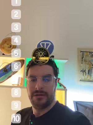 A man with a beard and glasses stands in front of a wall, a coin with the Cardano ADA logo is balanced on his head.