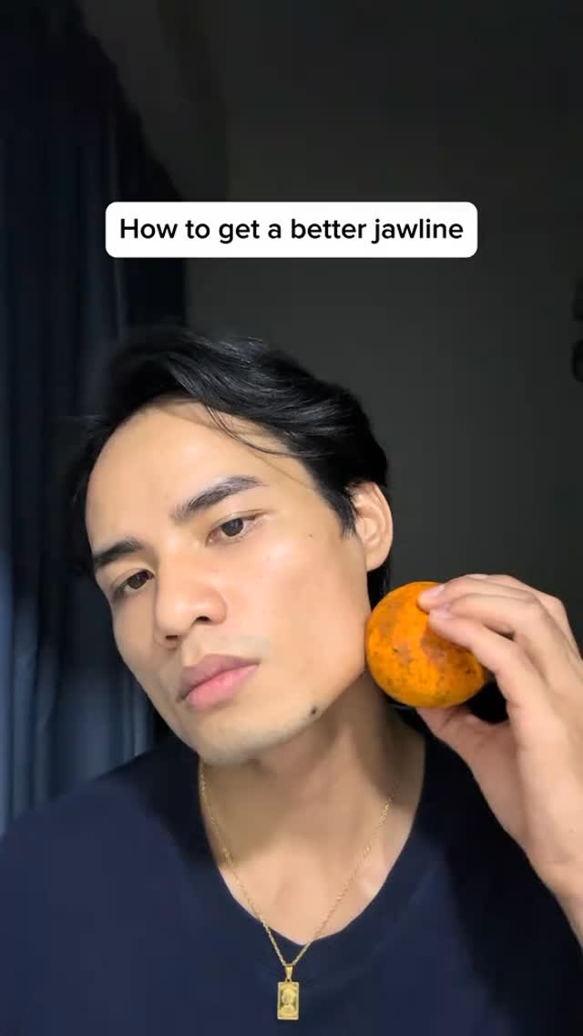A young man with dark hair and a dark blue shirt holds a round orange object up to his jawline, as if demonstrating a technique. Text overlay at the top reads, "How to get a better jawline."