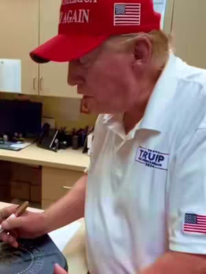 The cover image shows a man, presumably Donald Trump, wearing a red cap with the words "Make America Again" on it, and white shirt .The man is holding a pen and leaning over a document or object, appearing to be signing something. The background is a typical office setting.