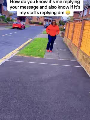 A woman in an orange sweater and dark blue pants walks on a sidewalk next to a grassy strip and a street. A red car is parked on the road in the background.