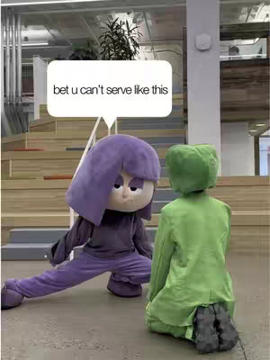 The cover image features two figures in costumes in what appears to be a building interior. One figure, dressed in purple, is in a crouching pose, seemingly posing, with a speech bubble above it. The other figure, dressed in green, is also kneeling facing the purple figure. The background includes stairs and architectural details.