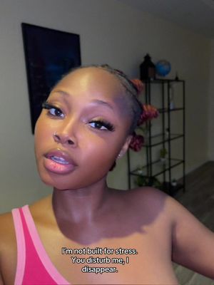 A selfie-style shot with a dark-skinned woman in the foreground looking directly at the camera. She has styled black hair, with a few red-tinted strands. The background is blurred but shows a room with a shelf holding decorative items and a framed artwork. The woman is wearing a pink tank top with a halter neck. Text overlayed at the bottom reads: 'I'm not built for stress. You disturb me, I disappear.'