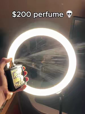 A hand holds a black perfume bottle with a gold design, spraying into a spotlight. The background is a dark room.