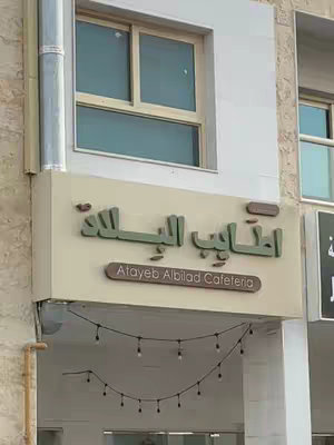 The image shows the facade of a building in an urban setting. The main focus is on a sign that reads "Atayeb Albilad Cafeteria" in both Arabic and English. The sign itself is mounted on a structure that extends from the building. Below the sign, a string of decorative lights are strung across the bottom. Above, there is a window with a wooden frame.