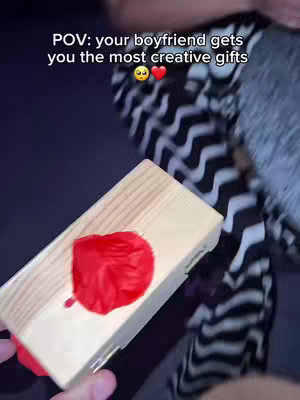 A person holding a wooden box with a red object on top. The person's clothing and the background are partially visible.