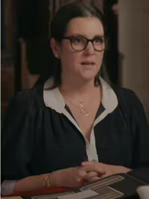 A medium shot of a woman wearing glasses and a black blouse with a white collar, sitting and looking slightly off-center. She has a gold necklace with a pendant, and a gold bracelet on her wrist. The background is blurred, suggesting an indoor setting, possibly an office or a room with wood paneling.