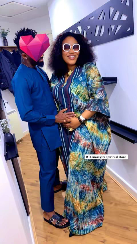 The image shows a couple posing intimately. The woman is wearing a patterned, colorful dress and sunglasses, while the man has his face obscured by a heart-shaped graphic. The background has a modern interior design with decorative elements, with the couple standing closely and touching hands.