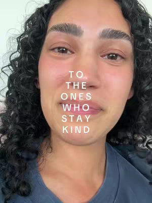 A close-up shot of a person with curly dark hair, facing the camera. The person has fair skin, visible eyebrows, and slightly parted lips. Text overlay in white, stacked vertically reads: TO THE ONES WHO STAY KIND.