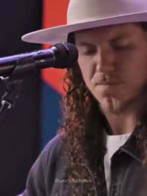A medium shot features a man with long, curly hair under a wide-brimmed hat, playing a guitar. A microphone is positioned in front of him. The background is blurred, with hints of color.
