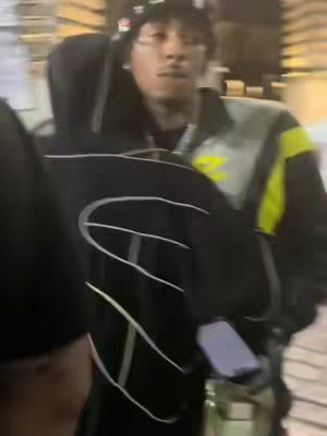 The image is a medium shot in a nighttime setting. A person, likely the creator, is the central figure, partially obscured by another person's back on the left, and is wearing a black Champion hoodie. They are looking at the camera. The background is blurred, suggesting motion or a crowded area. The lighting is dim, relying on ambient sources with a slightly warm/yellowish cast.