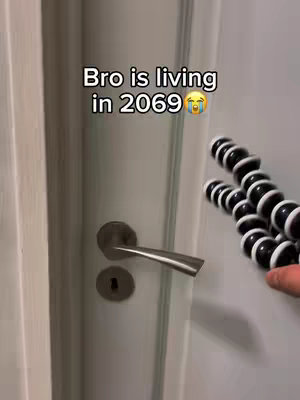 The image shows a white door with a silver handle and a keyhole. In the background, a black and white flexible tripod is positioned to the right, seemingly holding a phone. Text overlay in white font states, 'Bro is living in 2069', followed by a crying face emoji.