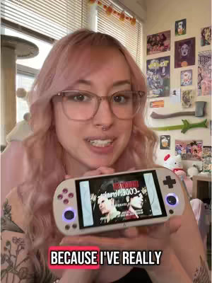 A young woman with pink hair, glasses, and a nose ring is holding a handheld gaming device. She is smiling and facing the camera, with the device held in front of her. The background is a room with various decorations and posters.