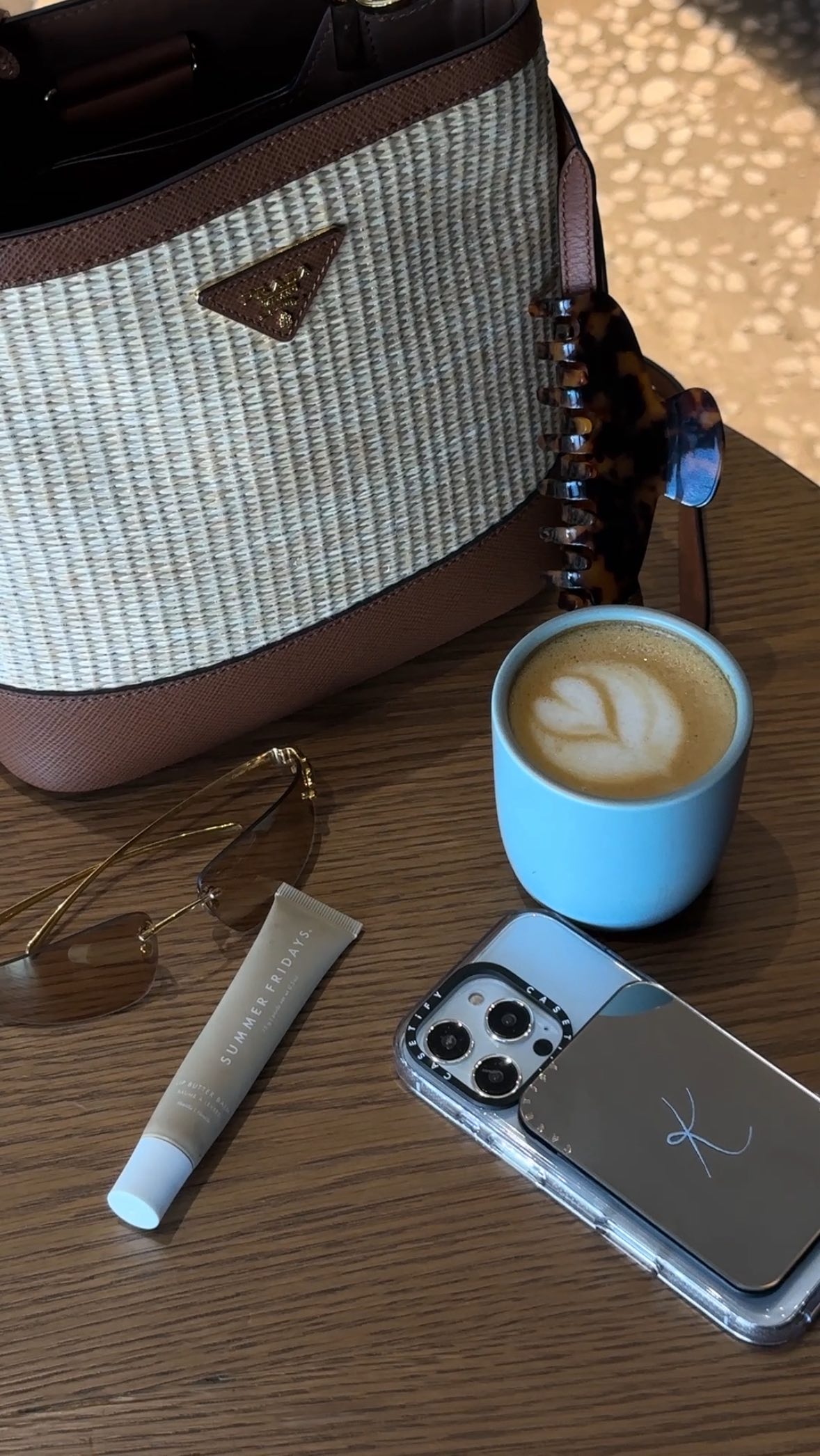 The image features a beautifully arranged collection of everyday items on a wooden surface. A woven handbag with a brown leather trim and a gold Prada logo is placed on the top left, to the right sits a tortoiseshell hairclip, a blue ceramic mug with a latte art design, a smart phone, a tube of `SUMMER FRIDAYS` lip balm, and a pair of sunglasses. The lighting appears to be natural, casting soft shadows creating an aesthetically appealing arrangement.