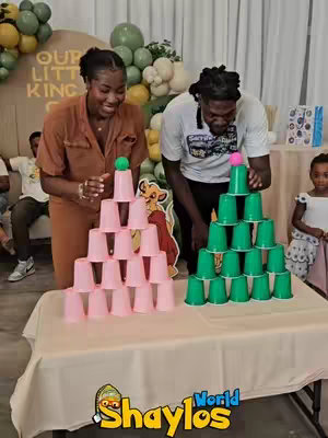 Two people, a woman and a man, are smiling while stacking cups in a game. The woman is wearing a brown jumpsuit, and the man has a white t-shirt and black hair. Behind them, there are party decorations and balloons. In the foreground, two stacks of cups are arranged on a table.