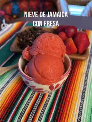 A close-up shot of a bowl of pink ice cream, with strawberries and a wooden tray in the background, all set on a colorful striped blanket.