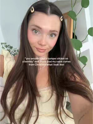 A selfie-style image featuring a young woman looking directly at the camera with a slightly upward gaze. She has long, dark brown hair styled with two golden hair clips, light makeup highlighting rosy cheeks and full lips. She is wearing a light-colored top. Text overlayed on the image provides additional context.