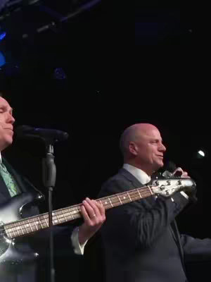 Two men performing on stage with a bass guitar and microphone. The image is dark with a spotlight on them.