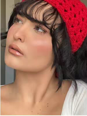 A selfie-style shot of a person looking up and to the left. They have dark hair with bangs. The person is wearing a bright red crocheted headband. Their skin has a rosy blush, and their lips are glossy. They are wearing a white top.