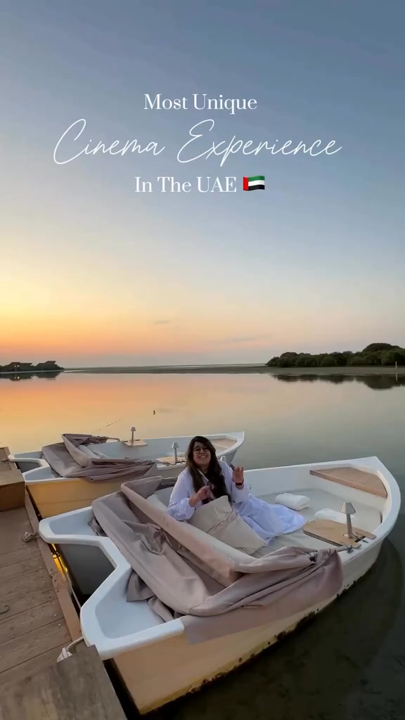 The image shows a woman sitting in a white boat on a calm body of water, with a backdrop of a sunset over a mangrove area. Text overlayed reads 'Most Unique Cinema Experience In The UAE' with the UAE flag. The boat has cream-colored cushions and a small table.