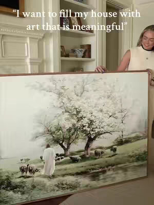 A woman holds up a large framed painting of a religious scene. The artwork depicts Jesus as a shepherd with sheep in a pastoral setting.