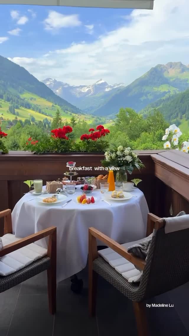 A scenic outdoor shot featuring a table set for breakfast on a balcony, overlooking a mountain range. The table is covered with a white tablecloth and set with plates, glasses, and food items like pastries and fruit. Red flowers are visible along the railing, and a backdrop of mountains under a blue sky.