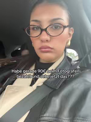 A selfie-style shot taken from inside a car. The focus is on a woman wearing glasses and a leather jacket. Text is overlaid at the bottom, and she appears to be looking at the camera with a slightly surprised expression. The overall composition is a close-up, centered on the woman's face.