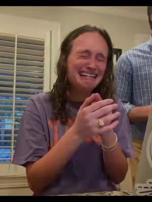 A woman with long brown hair is excitedly clapping her hands, looking up and to the left with tears in her eyes, in front of a window with a wooden frame and blinds. A man in a blue plaid shirt is seen in the background, and part of a kitchen.