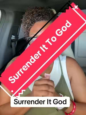 A woman in a car with a red banner overlaid with text 'Surrender It To God'. She has glasses and is wearing a white and green top, wrists with a red bracelet and a watch, hands clasped together. The background is out of focus, suggesting the car interior.
