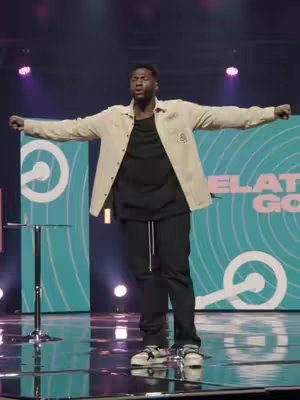 A man in a beige jacket and black pants stands on a reflective surface, arms outstretched. Behind him, a teal backdrop features white circles and the words "RELAT GOS". Stage lights illuminate the scene.