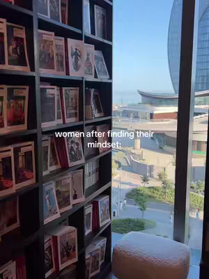A shot from inside a room with a large bookshelf on the left and a large window showing an office building and the sea in the background. The bookshelf is packed with books, and below it is a cushioned seat. The text overlay is placed over this bookshelf.