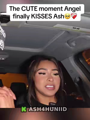 A woman is seated inside a car, looking towards the right with an expression that suggests she is speaking. The background indicates an outdoor setting likely at night with streetlights adding to the ambient light. The image is framed from a slightly low angle, possibly shot from the passenger seat. The woman has long lashes, light-colored skin, and light colored hair and a dark top.