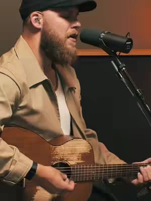 A man with a beard and a cap is playing an acoustic guitar and singing into a microphone. The scene is set in a studio with a muted background.