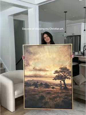 A woman holds up a framed painting. The painting depicts a landscape with a sunset or sunrise, a tree, and figures in the distance. The woman is smiling and standing in a well-lit room.