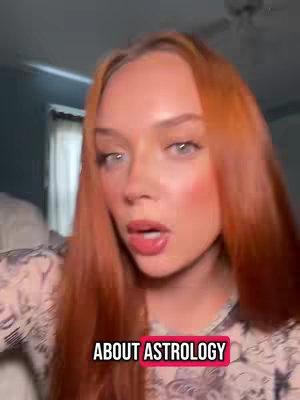 A selfie-style video cover image featuring a fair-skinned woman with long, auburn hair. She's looking directly at the camera with a slight open-mouth expression. She has light pink blush and her lips have a gloss, and her eyes are a light blue shade. The background is slightly blurred, but a glimpse of a room interior is visible. There is a text overlay over the image.