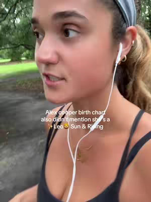 A young woman with light skin is seen from the chest up, wearing a dark tank top, a gray headband, and white earphones. Her dark hair is pulled back in a ponytail. She appears to be talking while looking slightly off-camera. Text overlaid on the image indicates 'Alex cooper birth chart also didn't mention she's a Leo Sun & Rising'. The background is blurred, suggesting an outdoor environment, perhaps a park, with visible green trees.