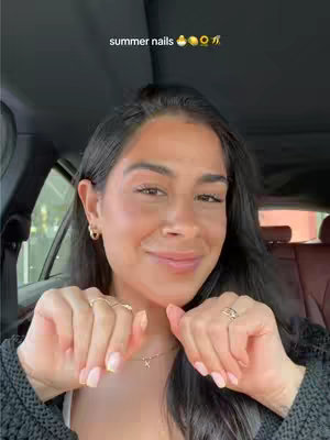A selfie-style shot taken inside a car. The background is dark, likely the inside of a vehicle, with the car's interior visible. The main subject is a woman with dark hair and visible earrings, smiling towards the camera. She's holding her hands up, showcasing her manicured nails, which are the central focus, and wearing a black top. Overlayed text, "summer nails", is present at the top of the image.