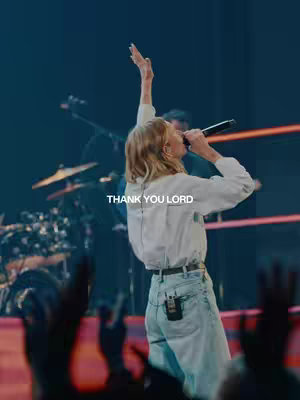 A live performance image captures a woman with blonde hair, holding a microphone and raising her arm towards the upper left, suggesting a moment of praise or worship. The background is blurred with stage lighting and a band setup, a text overlay at the bottom reads "THANK YOU LORD".
