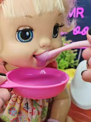 The cover image shows a close-up of a Baby Alive doll being fed with a pink spoon and bowl. The doll is in focus, with its open mouth, and a pink liquid coming from the spoon. The background is blurred, with hints of other colors and items.