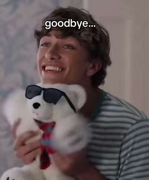 A young man with curly brown hair smiles widely, holding a white teddy bear wearing sunglasses and a red and black plaid tie. The man, dressed in a striped tee shirt, is in a well-lit indoor space. Text overlay 'goodbye...' appears in white against the background.