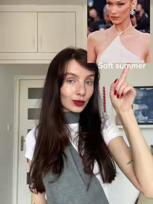A woman with long dark hair and red lipstick points her index finger up towards an image of Bella Hadid. The image is a casual indoor shot. The background includes a glimpse of doors, walls, and some upper cabinet doors.