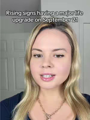 A woman with blonde hair is centered in the frame, gazing towards the camera with a subtle smile. Text overlayed at the top reads: "Rising signs having a major life upgrade on September 21". She wears a dark blue top and a delicate gold necklace. The background is a light, plain wall with a visible white door.