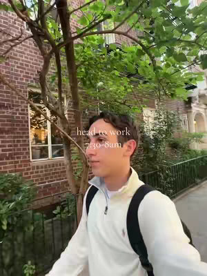 A young man walking on a sidewalk towards the camera, with a leafy tree overhead and a brick building in the background. Text overlay indicates "head to work 7:30-8am". The man is wearing a white quarter-zip sweater and a backpack.