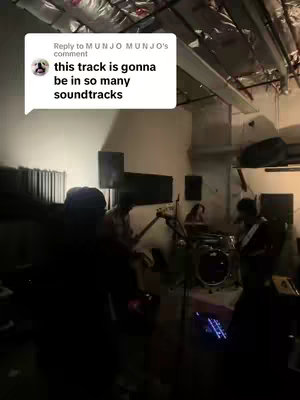 The image captures a moment from a music studio, filled with musicians playing their instruments. The band includes a guitarist, bassist, drummer, and a keyboardist. The lighting is dim, casting shadows throughout the space, and the walls are of a neutral color. A 'Reply' comment is shown at the top left corner of video frame.