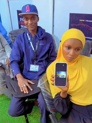 A medium shot features two individuals. On the left, a man in a blue cap and a matching blue jacket sits, holding a black tray. On the right, a woman in a yellow headscarf holds a smartphone displaying an app.