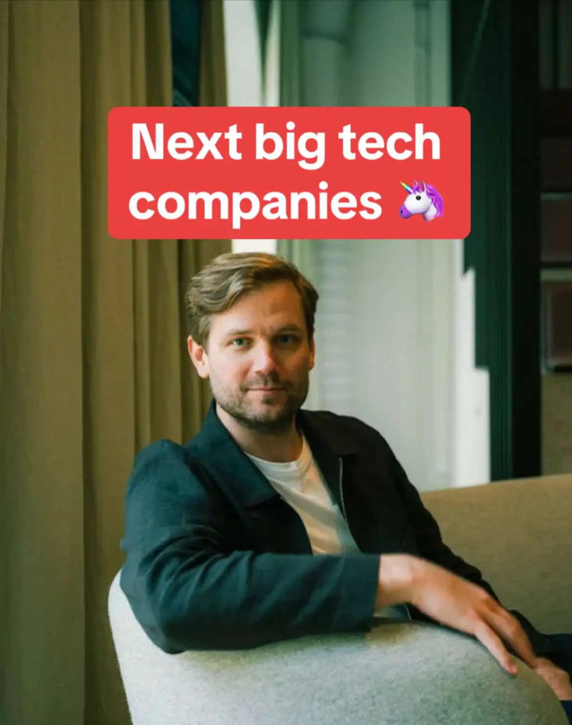 A man with short brown hair and a beard sits on a beige sofa. He is wearing a dark jacket over a white shirt. A red rectangular text box with the words "Next big tech companies" and a unicorn emoji is positioned above the man's head. The background is blurred with glimpses of a window and vertical paneled structure.
