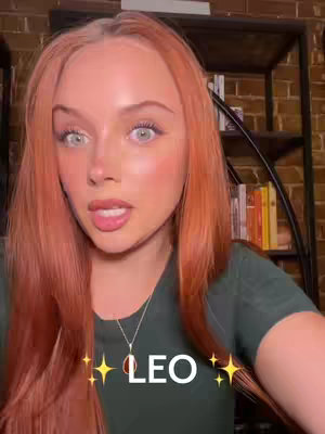 A young woman with reddish hair and fair skin is centered in the frame. She is looking at the camera and speaking. The background features a brick wall, bookshelves and warm lighting.